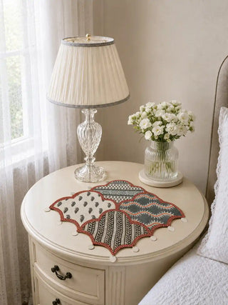 Riwaayat patchwork table mats with rust scalloped borders and hand-stitched embroidered patterns styled on a round white pedestal table with crystal lamp, fresh white flowers, and pearl bead embellishments in a serene bedroom setting.