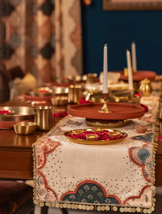 The Riwaayat table runner in cream with scalloped arch panels in teal and coral, scattered sparkle details and a round disc trim, on a dark wood dining table.