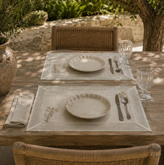 A pair of Rooted Grace embroidered placemats styled for an outdoor lunch on a weathered wood table with ceramic plates.