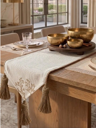 The Rooted Grace custom embroidered linen table runner styled beautifully on an oak table next to a decorative stack of raw brass serving bowls.