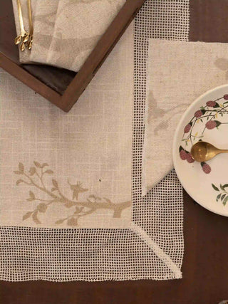 The Rooted Grace placemat in cream with a tonal branch motif and open-weave mesh border, styled on a dark wood table.