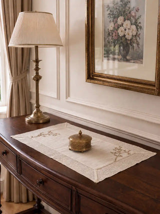 Rooted Grace cream linen table mats with sage botanical embroidered borders styled on a dark wood dresser with brass lamp, framed floral artwork, and decorative brass vessel in an elegant entryway setting.