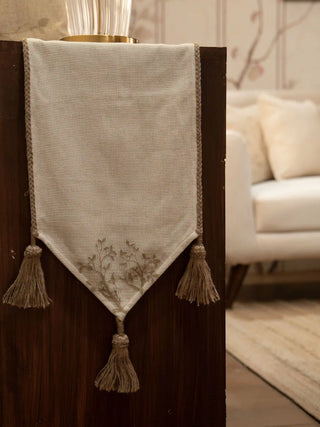 The Rooted Grace table runner in cream with a pointed end, three tan tassels, braided edging and a tonal branch motif, draped over dark wood furniture.