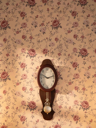 The Rose Heritage floral wallpaper installed on a wall, with trailing vines and buds in dusty rose and grey-green, behind a wooden pendulum clock.
