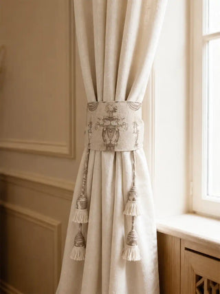 The Rosette Royale luxury embroidered cream tieback styled securely around a thick beige linen curtain panel next to a window.