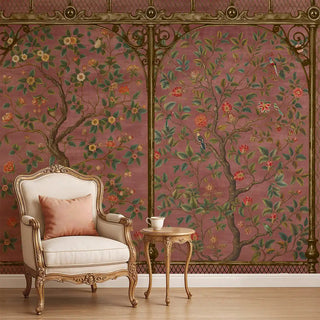 A dusty pink Chinoiserie wallpaper featuring ornate tree branches, blooming flowers, and green foliage framed with golden detailing, styled with a vintage armchair and side table.