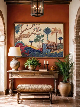 The Royal Jungle Safari wall art in a wooden frame on a terracotta limewash wall above a carved wooden console with a brass table lamp, white candles, potted plants, and a wicker bench in a Spanish-Indian heritage foyer