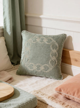 A luxury sage-green velvet cushion with white geometric lattice embroidery styled on a bed with light-colored linens and green curtains.