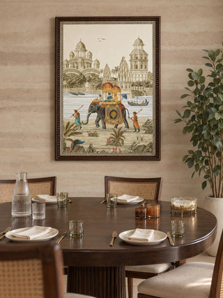 The Safar embroidered wall art in a beaded dark wood frame above a round walnut dining table set with cane-back chairs and ribbed glassware.