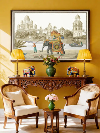 A large-scale Safar mural framed on a vibrant yellow wall above a carved wooden console and cane-backed armchairs