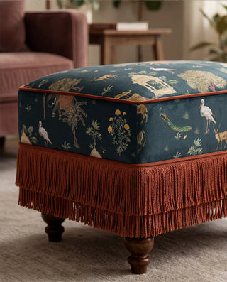 A detailed view of a Sair Blue upholstered ottoman featuring deep terracotta fringe and piping.