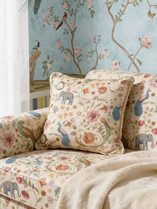 A luxury sofa and matching cushion upholstered in Sanjhi Cream fabric featuring hand-painted elephants and peacocks against a blue mural wall.