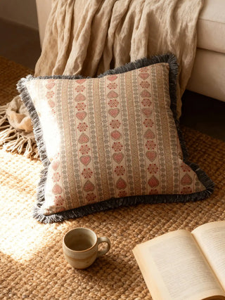 A luxury Sarangi cushion cover featuring terracotta floral bands and charcoal fringe resting on a textured jute rug near an open book.