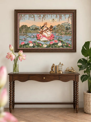 The Saraswati Maa wall art, framed and hung above a wooden console table styled with brass figurines and a potted lotus, showing Saraswati seated on a lotus playing the veena beside a swan.
