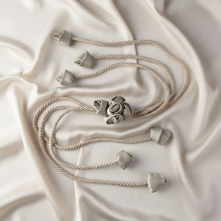 Flat lay of the Sculpted Petal Tieback showing the neutral braided cord, central rosette, and fabric bud charms on a satin background.