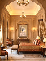 Shahenshah-e-Riyasat wall art in a gilt frame above a carved wooden console in a sandstone-arched bedroom with a brass chandelier and copper silk bedding
