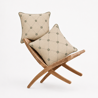 The Shahzar cushion set of 2 styled on a woven rattan bench, showing a diamond trellis pattern with embroidered coin medallions and pom-pom trim on natural linen.