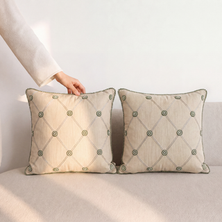 Pair of Shahzar hand-quilted ivory pillows with diamond quilting, brass studs, and beaded metallic trim edging displayed in natural light.