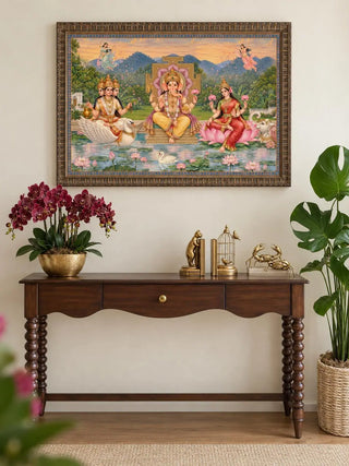 Framed Shree Lakshmi Ganesh Saraswati wall art in a carved bronze frame above a dark wood console with a brass bowl of burgundy orchids and gold accents