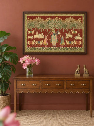 The Shrinathji Madhuban wall art in Maroon & Gold, framed and hung on a terracotta wall above a wooden console table styled with pink lotus flowers and brass figurines.