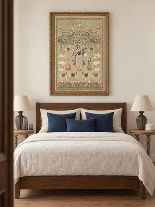 Shrinathji Madhuban portrait wall art in an ornate gold carved frame above a wooden bed with cream bedding and navy cushions, between round wooden bedside tables with grey fluted lamp bases.