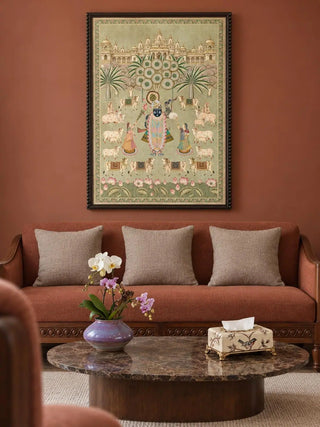 Shrinathji Madhuban portrait wall art in a dark beaded wood frame above a rust sofa with taupe cushions, beside a marble oval coffee table with an orchid in a purple glazed vase and a bird-print tissue box.