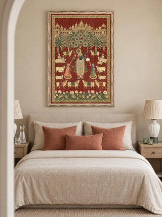The Shrinathji Madhuban wall art, portrait orientation, framed and hung above a beige upholstered bed with terracotta cushions and ribbed ceramic lamps.