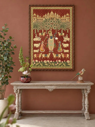 The Shrinathji Madhuban wall art, portrait orientation, framed and hung on a terracotta wall above a carved wooden console table styled with a potted lotus and a brass bird figurine.