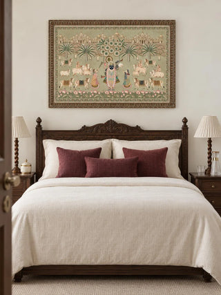 Framed Shrinathji Madhuban pista wall art above a carved dark wood bed with ivory bedding and maroon cushions in a bedroom