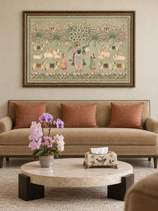 Framed Shrinathji Madhuban pista wall art hanging above a camel sofa with rust cushions and a round marble coffee table with pink orchids in a living room