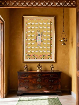 Shrinathji Pichwai wall art in a gilt frame above a dark walnut chest of drawers styled with three brass deity figurines and a hanging brass oil lamp in a warm saffron Indian interior with carved wooden ceiling frieze