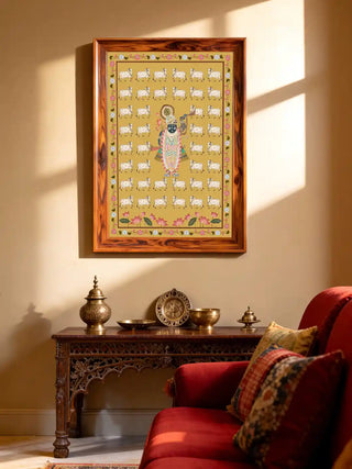 Shrinathji Pichwai wall art in a dark rosewood frame hung above a carved Indian console table with brass vessels, a decorative plate, and a red armchair with block-print cushion in a warm cream interior
