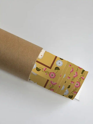 Shrinathji Pichwai wall art partially unrolled from its kraft paper shipping tube, revealing the saffron gold ground, pink floral vine border, and white sacred cow motifs