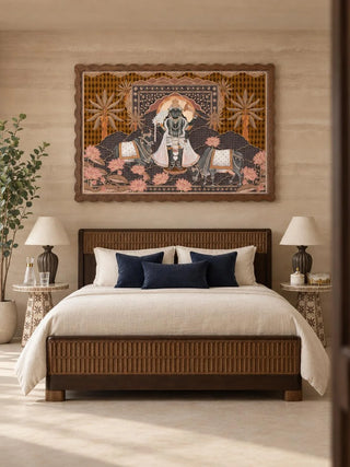 The Shyam Sundar embroidered wall art in a scalloped wooden frame above a woven-panel bed with navy cushions, between two ceramic table lamps.