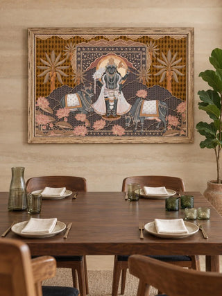 The Shyam Sundar embroidered wall art in a gilt wood frame above a walnut dining table with wooden chairs, white plates and green glassware.