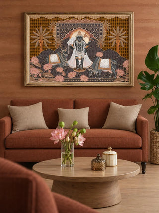 The Shyam Sundar embroidered wall art in a gilt frame above a rust sofa with beige cushions and a round wooden coffee table holding pink lotuses.