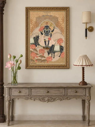 The Shyam Sundar wall art in Beige & Coral, in an ornate gold frame, above a carved grey-wood console table with a vase of pink lotus buds and a scallop-shade lamp.
