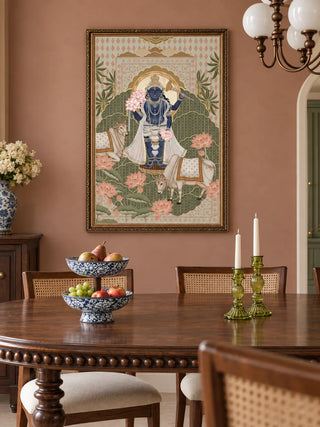 The Shyam Sundar wall art in Sage & Coral, in an ornate gold frame, on a dusty rose dining room wall above a round wooden table with a tiered blue-and-white fruit stand.