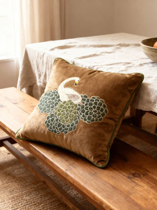 A luxury Silk Route cushion in golden-brown velvet featuring an embroidered swan and sage-green blossoms on a wooden bench.