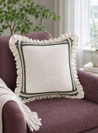 Spellbound ruffle cushion cover in dark green on a plum upholstered armchair with a fringed throw, beside a window in a living room.