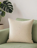 The Stardust off-white beaded linen cushion cover styled elegantly on a contemporary sage-green sofa panel next to a monstera plant.