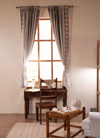 The Storah curtain in dove grey with a stacked diamond border and pinch-pleat header, hung on a brass rod at a tall wooden window.