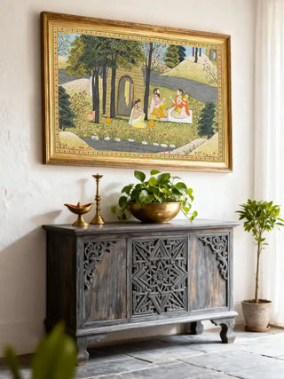 A framed Story of Rama mural styled above a hand-carved grey console table with brass accents and greenery.