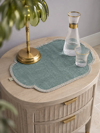 Sufra scalloped table mat in sage green styled on a light wooden nightstand with a brass lamp and glass carafe set.