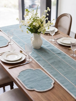 A close dining table view of the Sufra runner and mats set in Powder Blue with white plates, gold cutlery and a jug of white flowers.