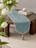 The Sufra table runner in Dusty Teal & Silver styled on a round wooden coffee table with a crystal vase of white tulips, stacked books and a candle, showing the scalloped edge and tasselled end.