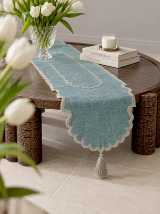 The Sufra table runner in Dusty Teal & Silver styled on a round wooden coffee table with a crystal vase of white tulips, stacked books and a candle, showing the scalloped edge and tasselled end.