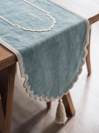 A close-up of the Sufra table runner in Powder Blue showing the hand-worked ari chain-stitch border and scalloped ivory trim with tassel.