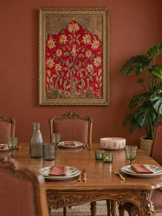 The Sultana wall art in an ornate gold frame on a terracotta wall above a carved wooden dining table with green glassware and rust napkins.