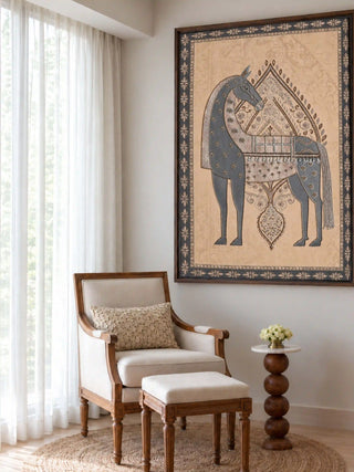 The Swarna Ashwa embroidered wall art framed above a cream armchair and footstool beside a turned wood side table and sheer curtains.
